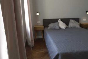 Ložnice po rekonstrukci s velkou postelí, jednoduchými nočními stolky a jemnými textiliemi u okna.