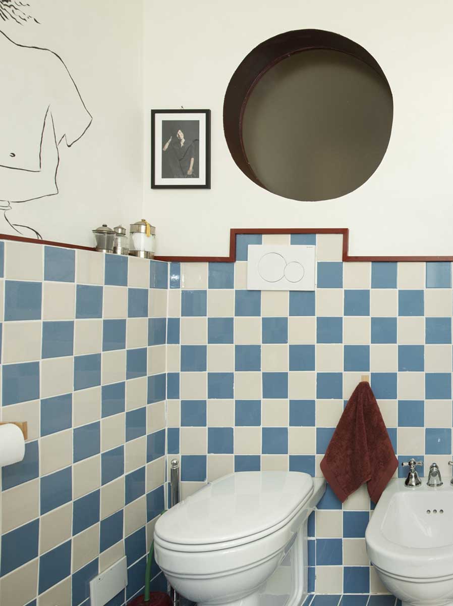 Toaleta s modrobílými čtvercovými obklady doplněná bidetem a kulatým okénkem ve stěně.