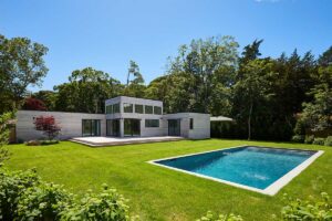 Moderní modulární dům s bazénem je zasazený do upravené zahrady plné zeleně a vzrostlých stromů.