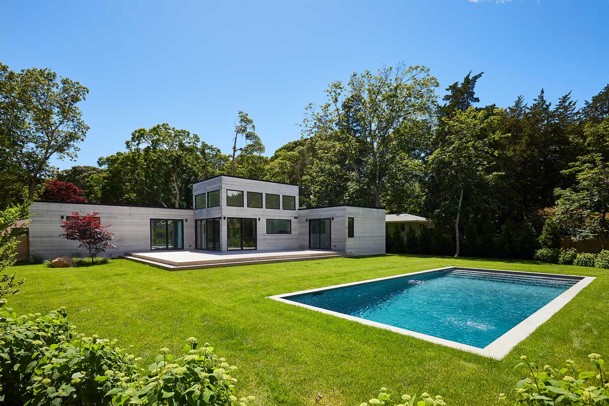 Moderní modulární dům s bazénem je zasazený do upravené zahrady plné zeleně a vzrostlých stromů.