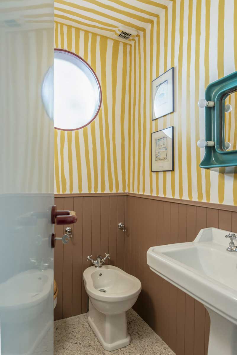 Malá koupelna s ručně malovanými žlutými pruhy na stěnách a kulatým oknem nad bidetem.