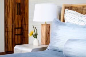 Ložnice v havajské vile s ratanovým čelem postele, dřevěnou lampou a jemnými přírodními textiliemi.