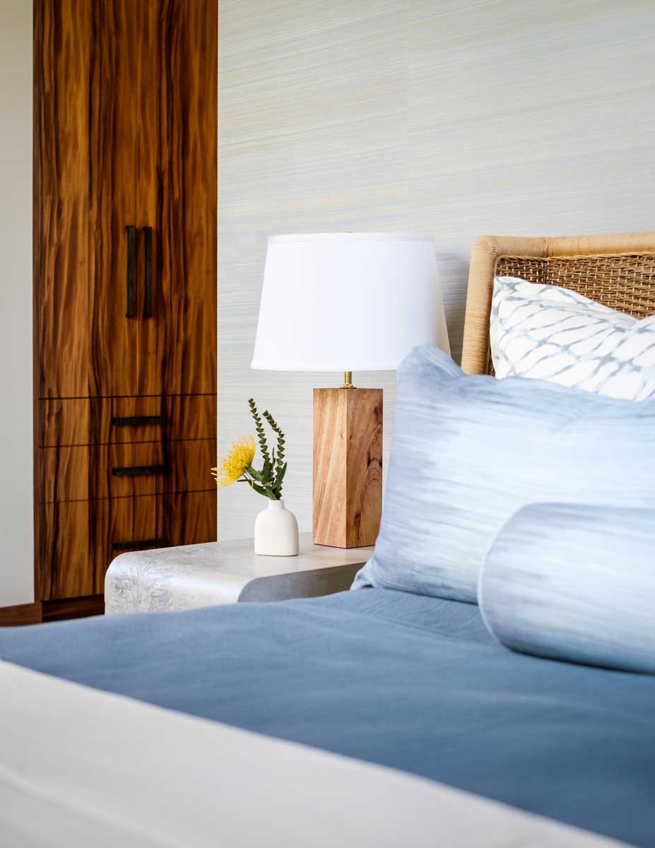 Ložnice v havajské vile s ratanovým čelem postele, dřevěnou lampou a jemnými přírodními textiliemi.