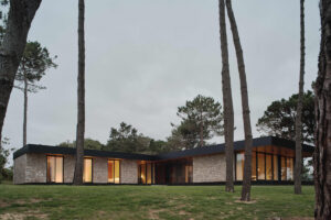 Nízký kamenný dům ve tvaru L stojí mezi stromy a citlivě zapadá do přírodního prostředí.