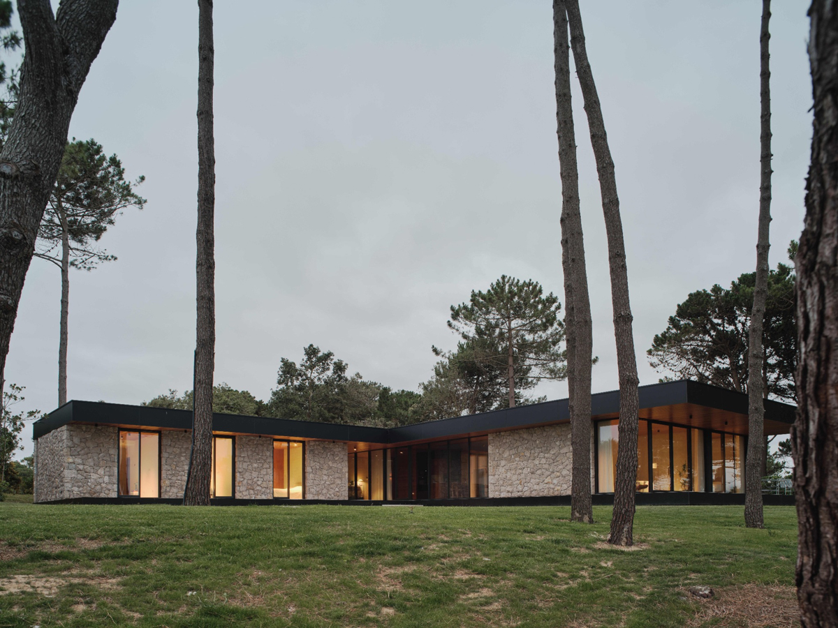 Nízký kamenný dům ve tvaru L stojí mezi stromy a citlivě zapadá do přírodního prostředí.