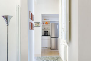 Dlouhá chodba se zachovanou terrazzo podlahou a vysokými okny vede do kuchyně v zadní části bytu.