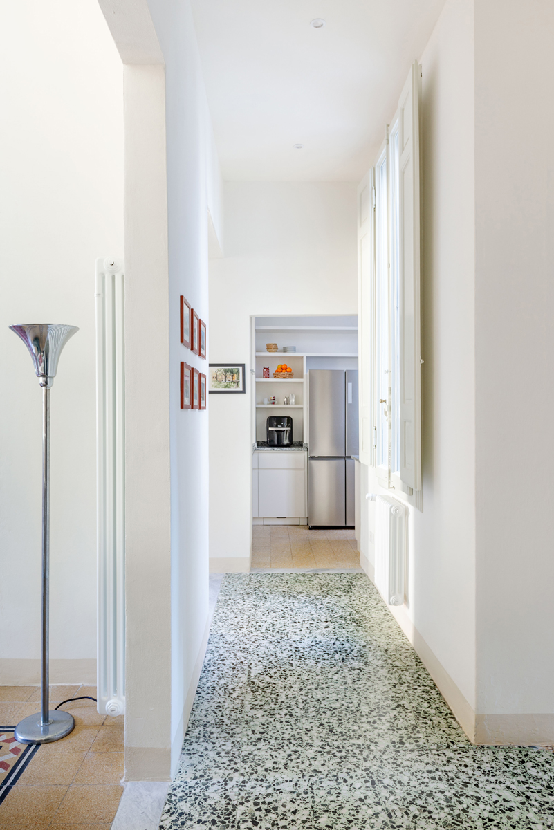 Dlouhá chodba se zachovanou terrazzo podlahou a vysokými okny vede do kuchyně v zadní části bytu.