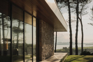 Zastřešený venkovní chodník podél prosklené fasády propojuje dům se zahradou a okolní krajinou.