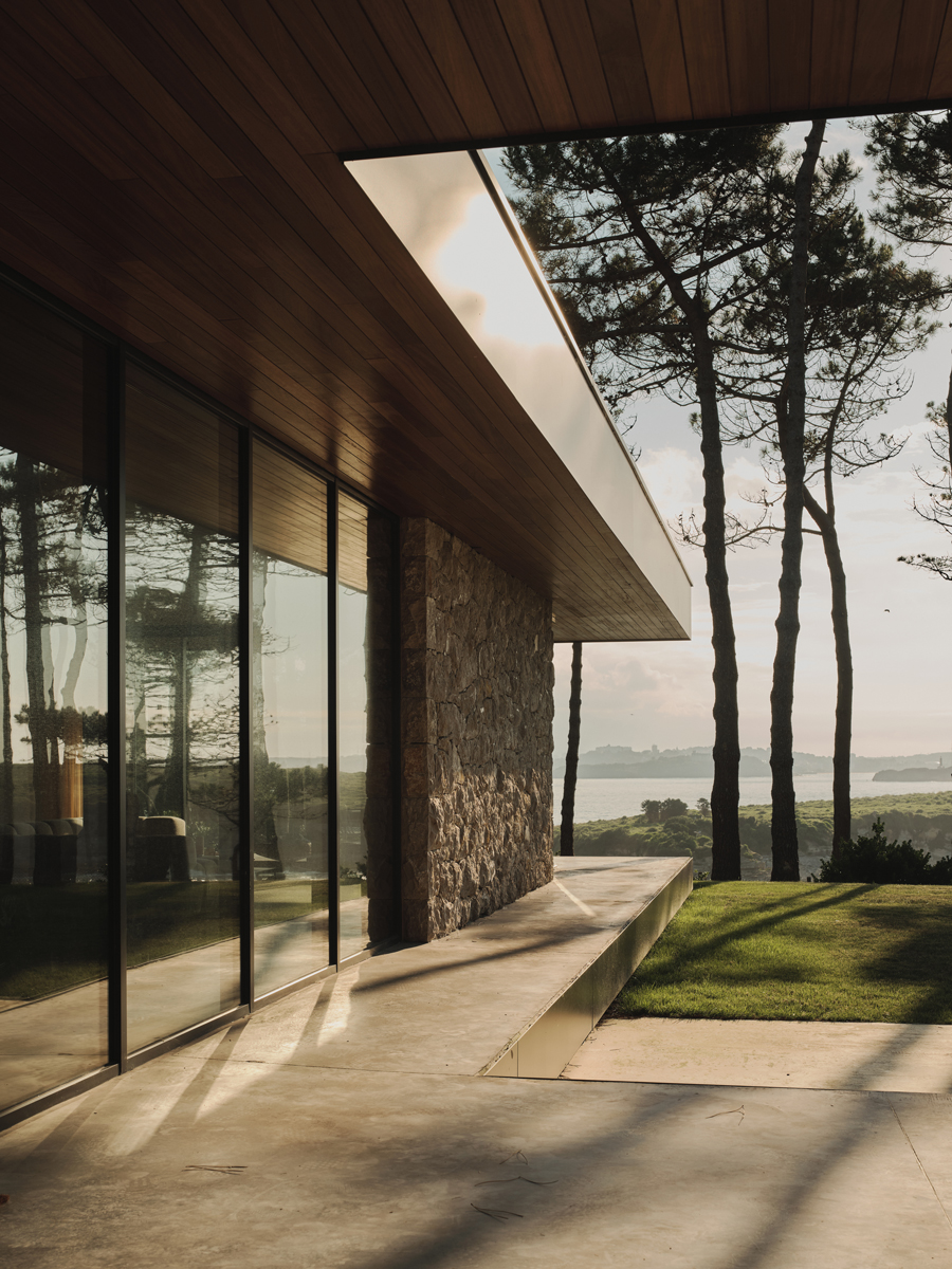 Zastřešený venkovní chodník podél prosklené fasády propojuje dům se zahradou a okolní krajinou.