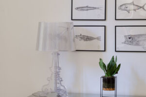 Vintage komoda se skleněnou deskou, transparentní lampou a kolekcí obrazů ryb vytváří stylový detail interiéru.