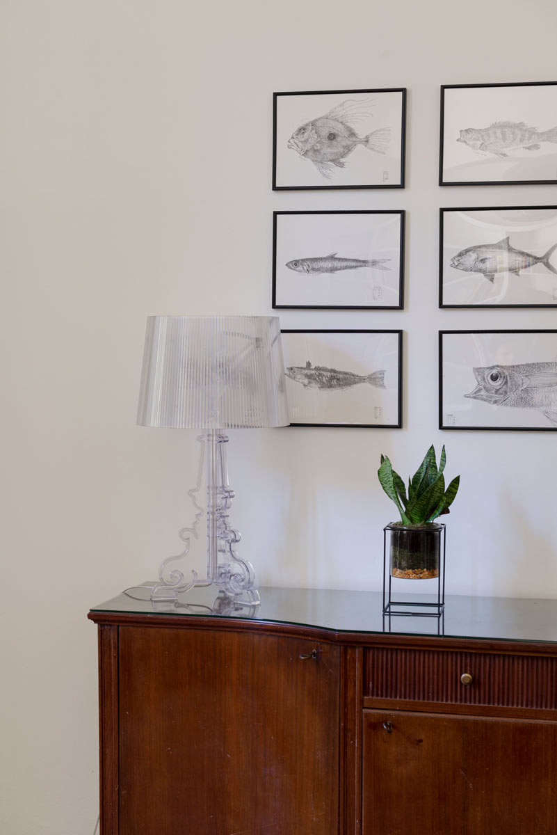 Vintage komoda se skleněnou deskou, transparentní lampou a kolekcí obrazů ryb vytváří stylový detail interiéru.