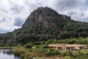 Dům zasazený v krajině u vodní plochy s výhledem na lesnatý svah.