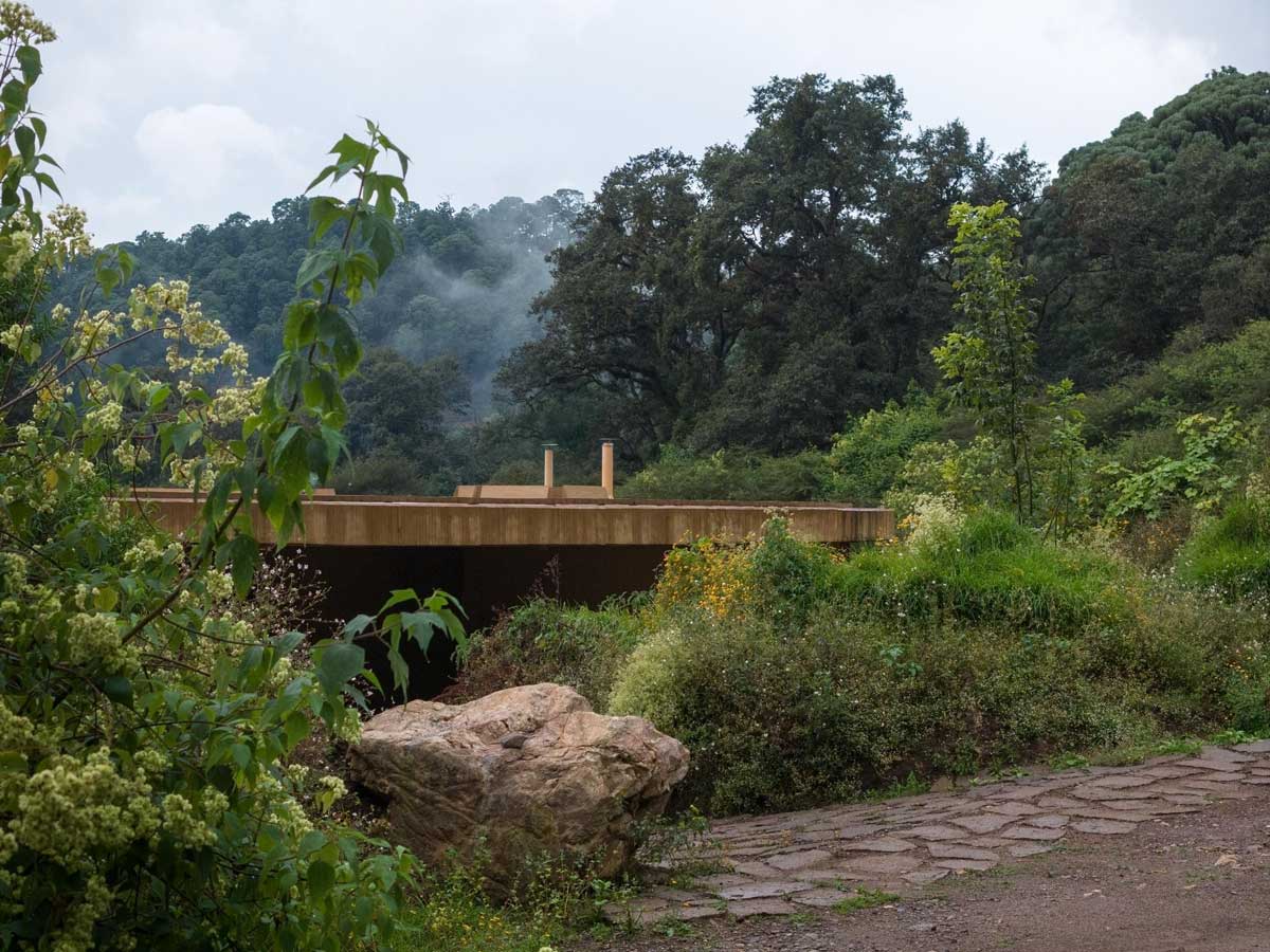 Nízký kruhový dům částečně zapuštěný do terénu obklopený divokou vegetací.