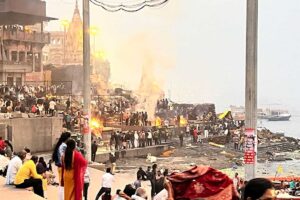 Spalovací rituály na břehu řeky Gangy s davem lidí a kouřem stoupajícím do vzduchu.