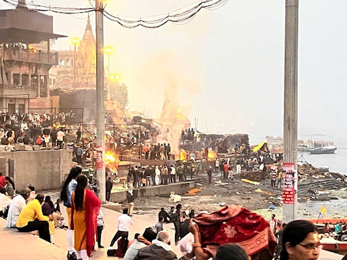 Spalovací rituály na břehu řeky Gangy s davem lidí a kouřem stoupajícím do vzduchu.