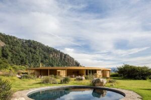 Kruhový dům zasazený do krajiny s malým jezírkem v popředí a zelenými kopci v pozadí.