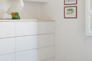 Bílá komoda na subtilních nožkách doplněná dekorativní lampou a grafickými obrazy zdobí klidnou část bytu.