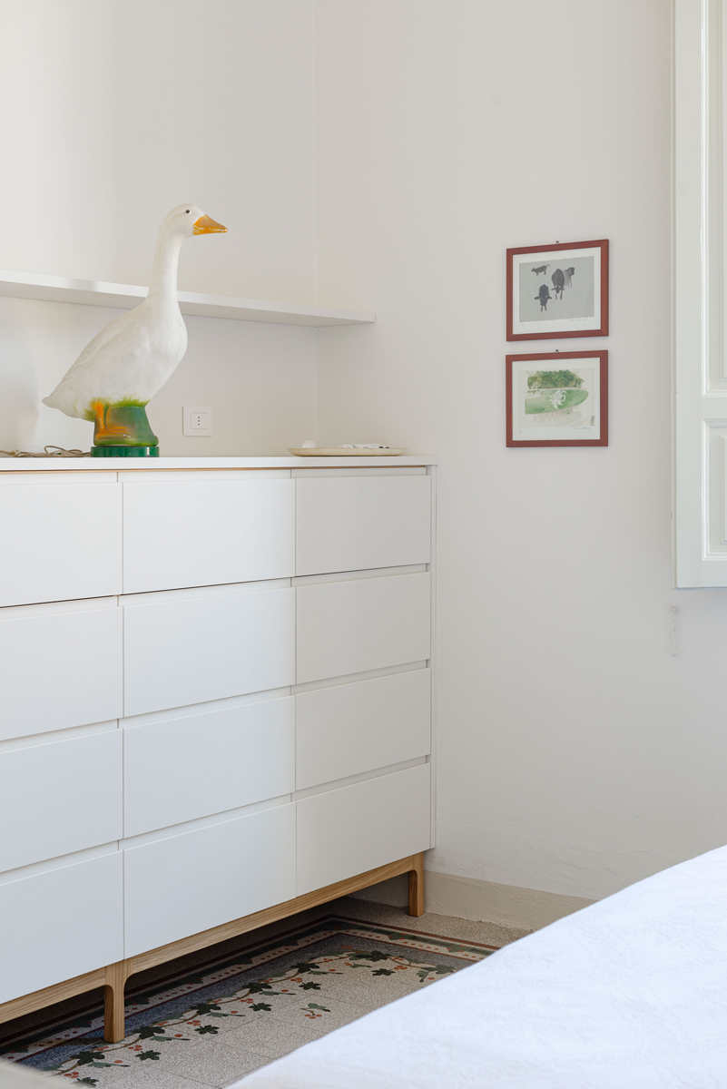 Bílá komoda na subtilních nožkách doplněná dekorativní lampou a grafickými obrazy zdobí klidnou část bytu.