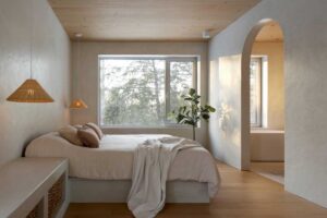 Minimalistická ložnice s dřevěným stropem, jemnými textiliemi a velkým oknem propojujícím interiér s lesní krajinou
