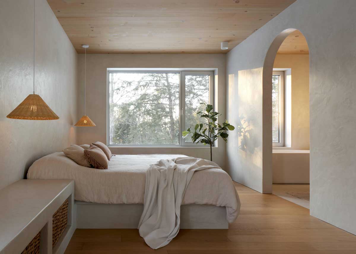 Minimalistická ložnice s dřevěným stropem, jemnými textiliemi a velkým oknem propojujícím interiér s lesní krajinou