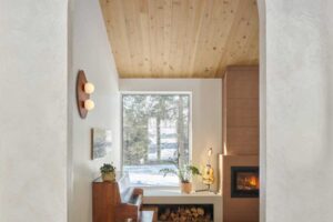 Pohled skrz obloukový otvor do obývacího prostoru s krbem, klavírem a velkým oknem do lesa