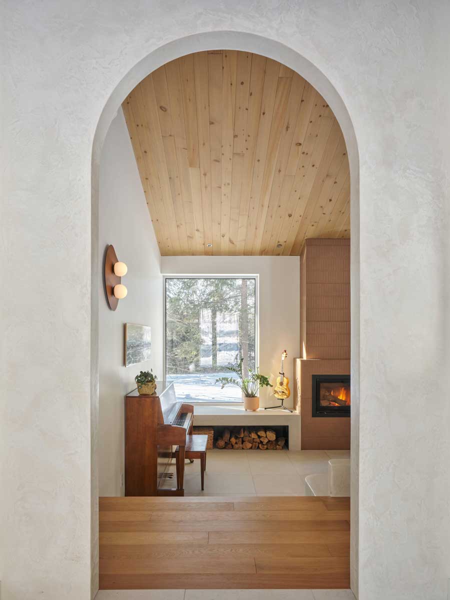 Pohled skrz obloukový otvor do obývacího prostoru s krbem, klavírem a velkým oknem do lesa