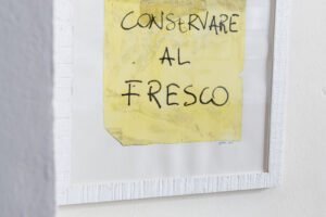 Detail nástěnného obrazu s italským nápisem doplněný skleněnou lampou a minimalistickým nábytkem v jídelní části bytu.