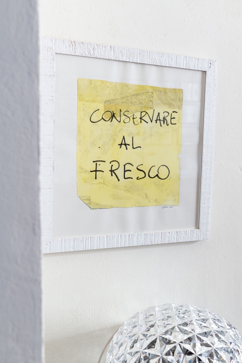 Detail nástěnného obrazu s italským nápisem doplněný skleněnou lampou a minimalistickým nábytkem v jídelní části bytu.