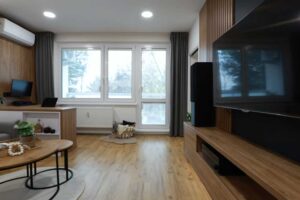 Světlý obývací pokoj s dřevěnou podlahou televizní stěnou a velkým oknem působí otevřeně a vzdušně.