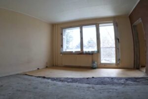 Původní obývací pokoj v panelákovém bytě s opotřebovanou podlahou a prázdným prostorem působí chladně a neútulně.