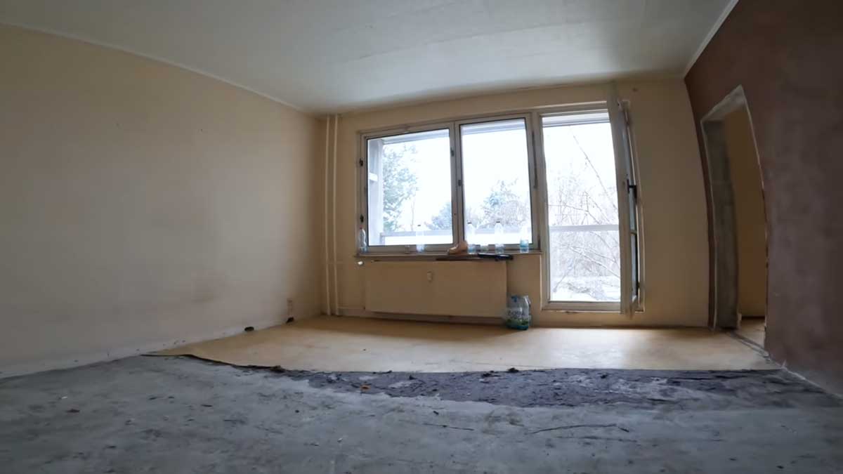 Původní obývací pokoj v panelákovém bytě s opotřebovanou podlahou a prázdným prostorem působí chladně a neútulně.