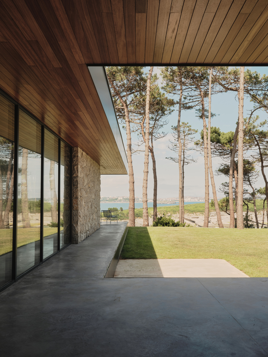 Krytá terasa a kamenná stěna rámují výhled do zahrady a směrem k moři mezi vysokými stromy.
