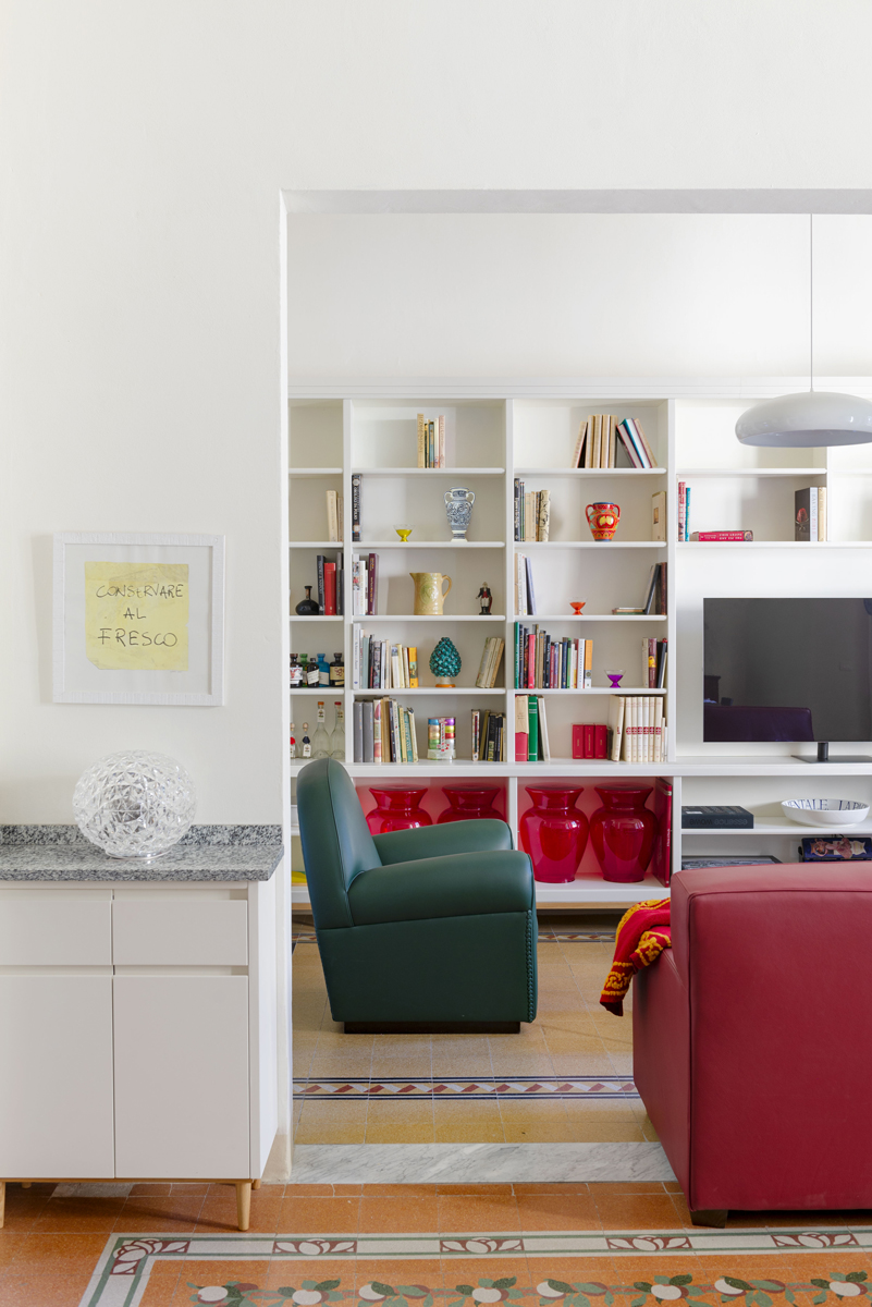 Otevřený pohled z jídelny do obývacího pokoje odhaluje vestavěnou knihovnu, zelené křeslo a výraznou červenou pohovku.