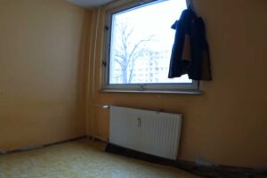 Původní pokoj s linoleem a jednoduchým oknem působí zastarale a postrádá jakýkoliv designový charakter.