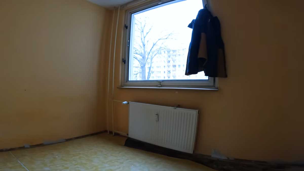 Původní pokoj s linoleem a jednoduchým oknem působí zastarale a postrádá jakýkoliv designový charakter.