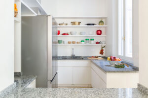 Žulová pracovní deska rámuje světlou kuchyň s otevřenými policemi, ovocem a pečlivě uspořádaným nádobím.