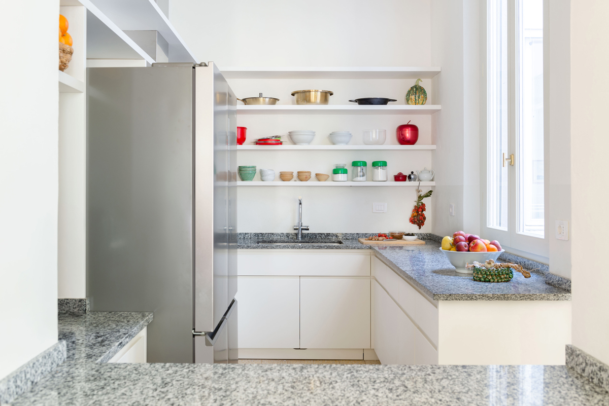 Žulová pracovní deska rámuje světlou kuchyň s otevřenými policemi, ovocem a pečlivě uspořádaným nádobím.