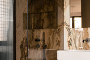 Koupelna v béžových tónech kombinuje mramorové obklady s tmavými prvky, čímž vzniká vyvážený a harmonický celek.