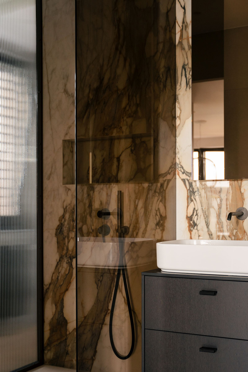 Koupelna v béžových tónech kombinuje mramorové obklady s tmavými prvky, čímž vzniká vyvážený a harmonický celek.