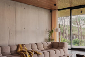 Prostorný obývací pokoj s měkkou sedací soupravou, betonovou stěnou a výhledem do zeleně působí útulně.