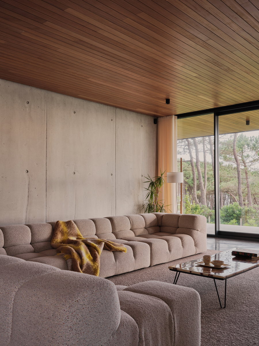 Prostorný obývací pokoj s měkkou sedací soupravou, betonovou stěnou a výhledem do zeleně působí útulně.