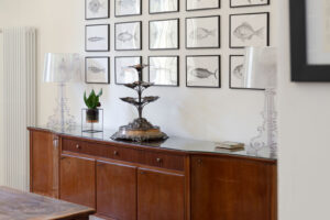 Vintage komoda s transparentními lampami a kolekcí kreseb ryb vyniká na pozadí vysoké světlé stěny.