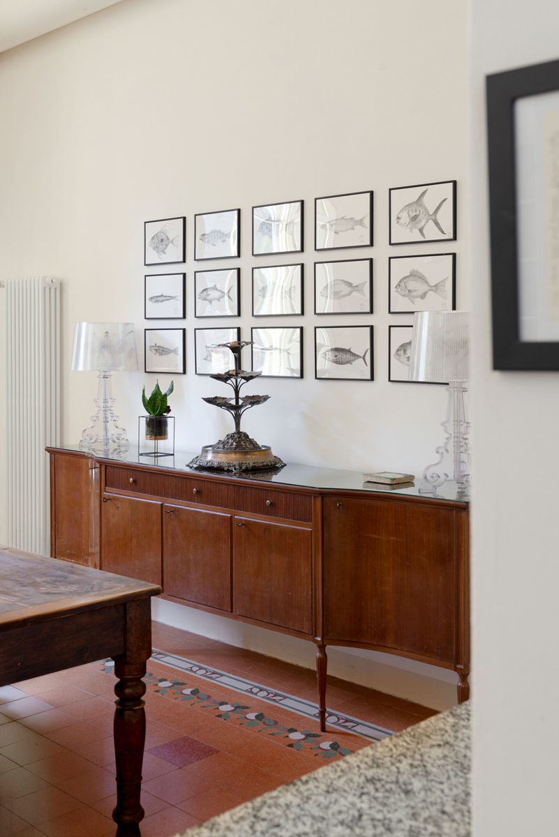 Vintage komoda s transparentními lampami a kolekcí kreseb ryb vyniká na pozadí vysoké světlé stěny.