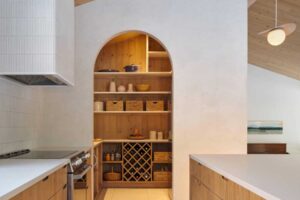 Kuchyň s dřevěnou linkou a obloukovým vstupem do spíže s otevřenými policemi a úložnými koši