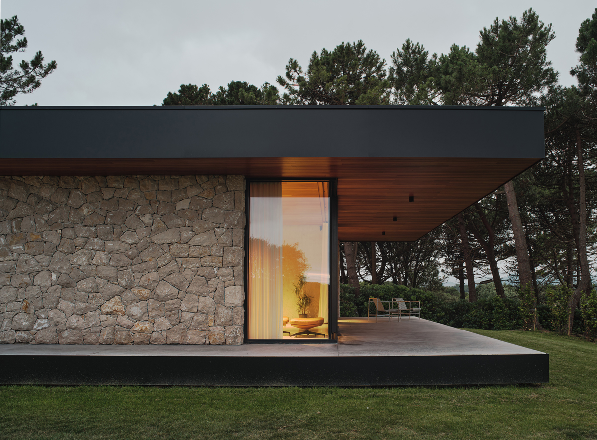 Večerní pohled na prosklený roh domu ukazuje osvětlený interiér a krytou terasu obrácenou do zahrady.