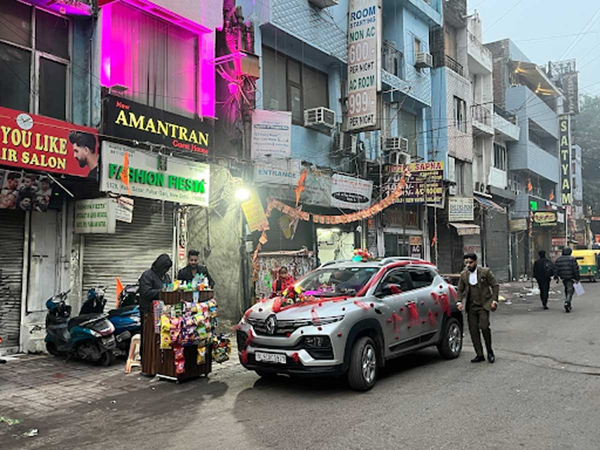 Ulice v Dillí s obchody, neonovými světly a ozdobeným autem projíždějícím městem.