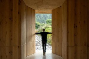 Průchod z interiéru do krajiny s dřevěnými stěnami a siluetou postavy v pohybu.