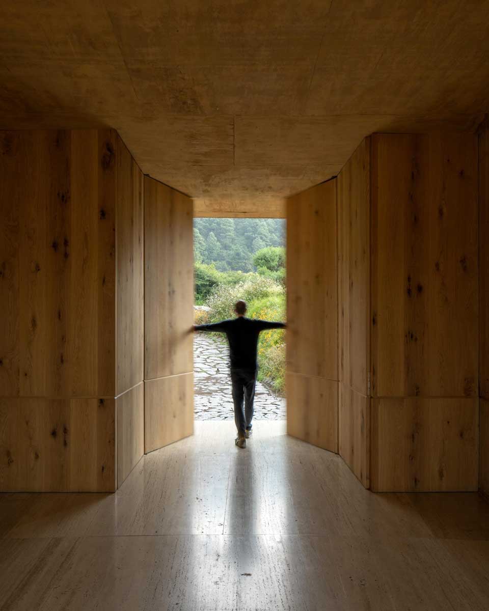 Průchod z interiéru do krajiny s dřevěnými stěnami a siluetou postavy v pohybu.