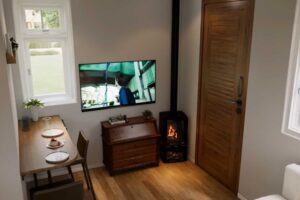 Obytná část tiny house s jídelním stolem, televizí a krbovými kamny u vstupu.