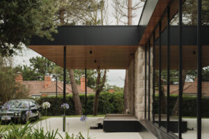 Vstupní část domu s dlouhým přesahem střechy, prosklenou fasádou a upravenou zahradou působí elegantně a klidně.